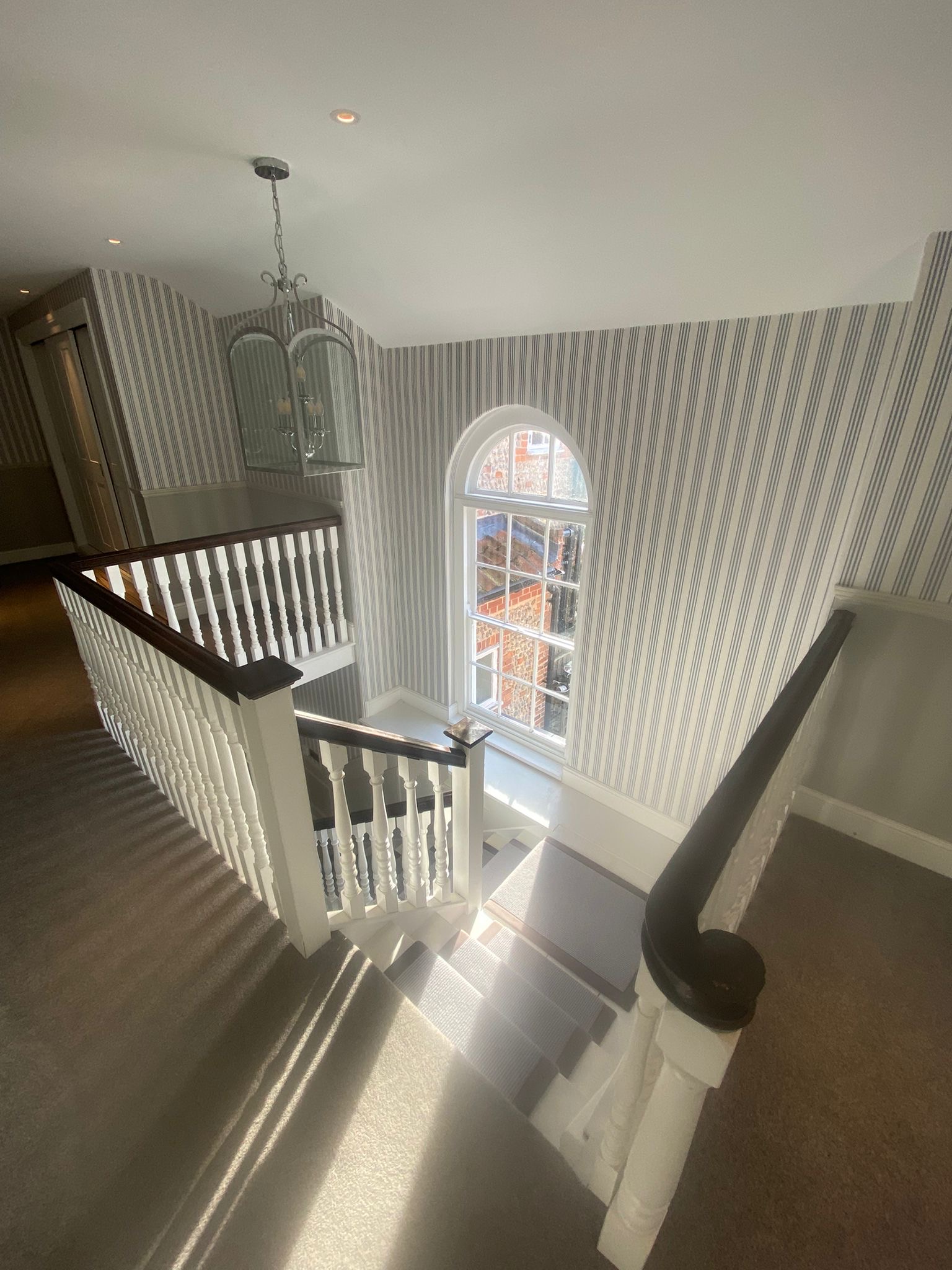 Stair landing with striped wallpaper, white balustrade and an arched window, decorated by Bywater